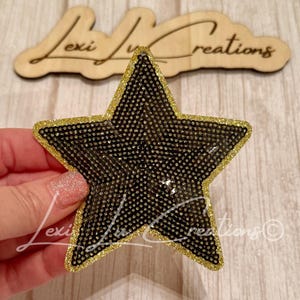 May include: Black star-shaped embellishment with gold glitter trim. The star is covered in small, shiny black beads. The background includes a wooden sign with the text "Lexi Lux Creations".