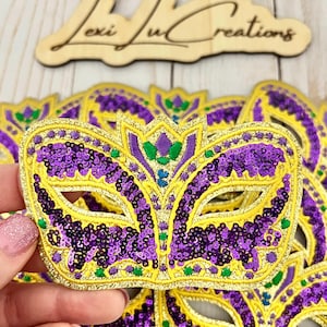 May include: A set of purple, gold, and green Mardi Gras mask-shaped iron-on patches. The masks are decorated with sequins.