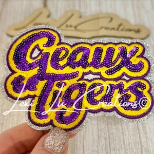 May include: A decorative patch with the words "Geaux Tigers" in purple and yellow, outlined with silver glitter. The patch is embroidered with sequins and has a cursive signature at the bottom.