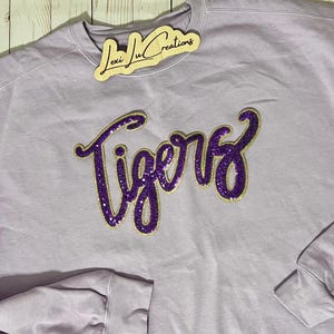 May include: A lavender sweatshirt with a purple and gold sequined "Tigers" design.