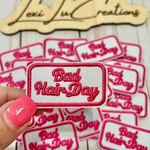 Pode incluir: Uma coleção de patches retangulares brancos com uma borda bordada rosa. Cada patch apresenta a frase "Bad Hair Day" em script rosa. Os patches estão empilhados, com um sendo segurado por uma mão com esmalte rosa.