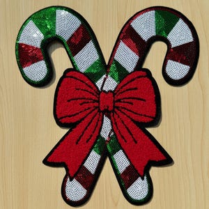 May include: A festive Christmas decoration featuring two candy canes with red, white, and green sequins. A large red bow is centered between the candy canes. The candy canes have a black outline.