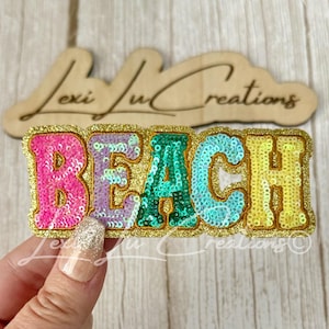 May include: A patch featuring the word BEACH in multi-colored sequins with a gold glitter border. Each letter is a different color: pink, lavender, green, light blue, and yellow. The patch is positioned in front of a wooden sign with the text Lexi Lu Creations.
