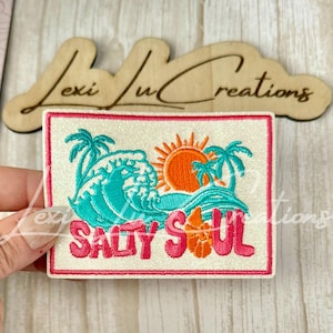 May include: A rectangular embroidered patch with a white glitter background and a pink border. The design features a teal ocean wave, an orange sun, palm trees, and an orange seashell. The text SALTY SOUL is embroidered in pink letters.