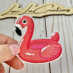 May include: Embroidered patch of a pink flamingo float. The flamingo has a black beak and eye, and a white border. The patch is held in a hand.