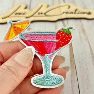 Peut inclure: Écusson brodé représentant un cocktail avec une garniture de fraise et un petit parasol. La boisson est rose et rouge, avec un bord et une tige bleus. L'écusson est tenu dans une main.