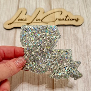 May include: A silver sequined Louisiana state-shaped embellishment. The sparkly decoration is held in a hand, with a wooden sign that reads "Lexi Lu Creations" in the background.