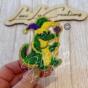 May include: A glittery alligator-shaped Mardi Gras pin. The alligator is green and yellow with purple accents, wearing a jester hat and holding a scepter. The pin is embellished with sequins and glitter.