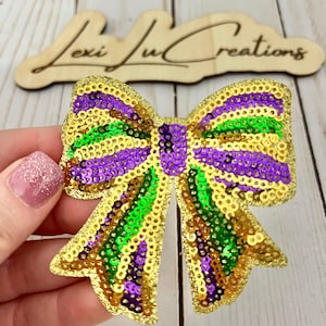 May include: A sequined bow in gold, purple, and green, with a Mardi Gras theme. The bow is held by a hand with a glittery pink nail. The wooden sign in the background says "Lexi Ju Creations".