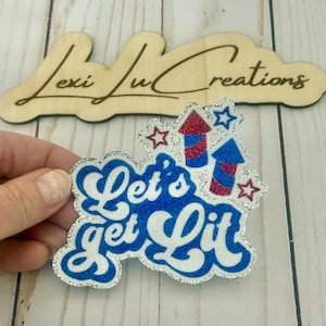 May include: A patriotic-themed pin with the phrase "Let's get Lit" in white, blue, and silver glitter. The pin features red and blue fireworks and star accents, perfect for celebrating Independence Day.