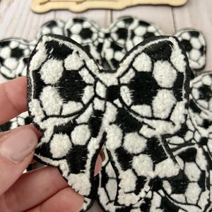 May include: A set of black and white soccer ball-themed fabric bows. The bows are made of a soft, plush fabric and feature a soccer ball pattern.