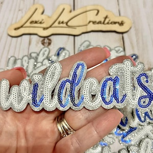 May include: A sequined "Wildcats" word art piece in silver and blue. The word is in a cursive font, with silver glitter outlining the letters and blue sequins filling the letters. The background is blurred, with other similar pieces visible.