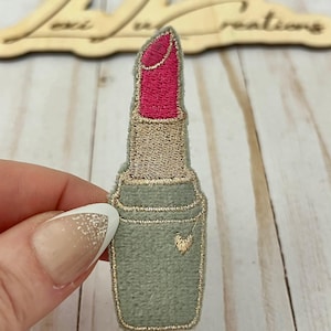 May include: Embroidered lipstick patch with a pink lipstick and a gold outline. The lipstick has a light green base with a small gold heart detail. The patch is held by a person.