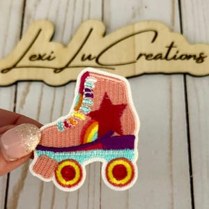 May include: Embroidered patch of a roller skate in shades of pink, red, yellow, and purple. The skate features a red star and a rainbow design. The patch is held in front of a wooden sign that reads "Lexi Lu Creations".