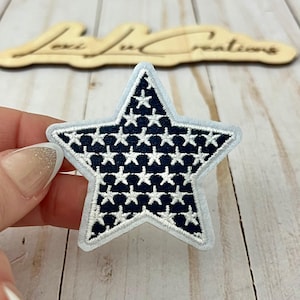 May include: Embroidered star patch with a navy blue background and white stars. The patch has a light blue border. The patch is held by a hand. The background includes a wooden sign that says "Lexi Lu Creations".