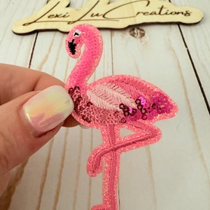 May include: Pink flamingo sequin patch with glitter. The flamingo is standing on one leg with its head turned to the side.