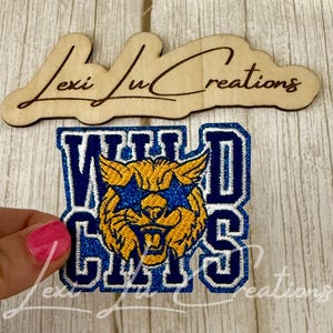 May include: A blue and gold embroidered patch with the words "Wild Cats" and a wildcat mascot. The patch is held by a person with pink nail polish. A wooden sign with the text "Lexi Lu Creations" is above the patch.