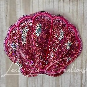 May include: A pink sequined seashell-shaped applique with a dark pink embroidered border. The shell is filled with small, shiny sequins in shades of pink and silver. The text "Lexi's Creations" is visible on the bottom left.