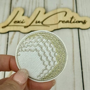 May include: Embroidered golf ball patch with white and gold thread. The patch is round and features the textured pattern of a golf ball. The background includes a wooden sign that reads "Lexi Lu Creations."