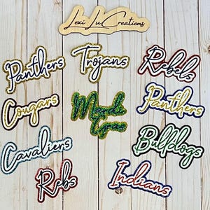 May include: A collection of 11 glittery iron-on patches with various school mascot names, including "Panthers", "Trojans", "Rebels", "Cougars", "Mudcats", "Bulldogs", "Indians", and "Cavaliers".