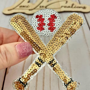 May include: A baseball-themed embellishment featuring a white baseball with red stitching and the number "11", crossed gold sequined bats, and silver sequin trim. The design is likely for crafts or accessories.