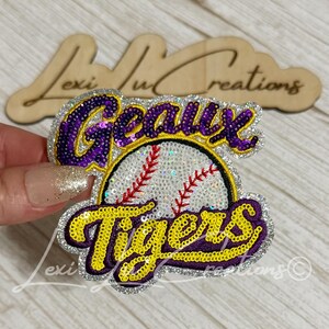 May include: A sequined patch featuring a white baseball with red stitching in the center. The word Geaux is embroidered in purple with a yellow outline above the ball, and the word Tigers is in yellow with a purple outline below the ball.
