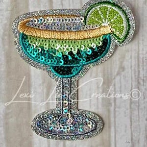 May include: A sequined margarita glass applique with a lime wedge. The glass features a gradient of teal, green, and gold sequins, with a silver glitter border. The design is a fun, festive accessory.