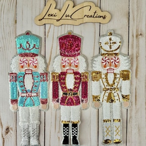 May include: Three sequined nutcracker ornaments in teal, red, and gold. Each has a white face with a candy-cane-like design for eyes and mouth. The ornaments are embellished with silver and gold sequins and are on a light wood background.