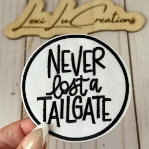 May include: A white circular patch with a black border and the phrase "NEVER lost a TAILGATE" in black script. The patch is held in a hand, with a wooden sign in the background that reads "Lexi Lu Creations".