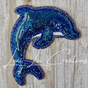 May include: A blue sequined dolphin patch with a white belly and a dark blue outline. The patch is in the shape of a dolphin jumping out of the water. The patch is on a light gray, textured surface.