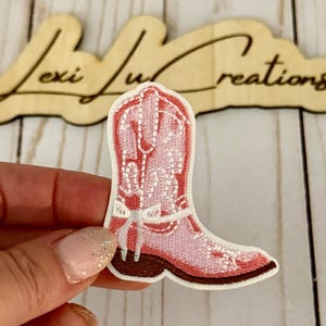 May include: A pink embroidered cowboy boot patch with white floral details and a brown sole. The patch is held in front of a wooden sign that reads "Lexi Lu Creations".
