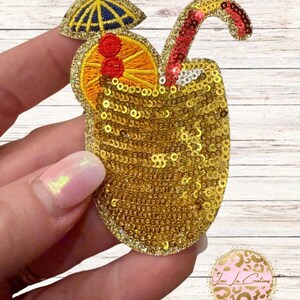 May include: A cocktail-shaped embroidered patch featuring gold sequins covering the glass, an orange slice with two red cherries, a blue and yellow cocktail umbrella, and a red sequined straw extending from the top of the drink.