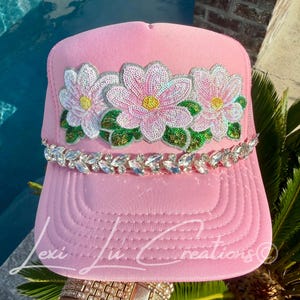 May include: A light pink trucker hat featuring a sequined floral applique with three pink flowers, yellow centers, and green leaves. A decorative band of clear faceted crystals sits across the base of the crown above the brim. Text reads Lexi Lu Creations.
