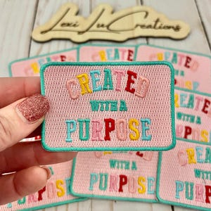 May include: Pink embroidered patch with the words "CREATED WITH A PURPOSE" in colorful letters. The patch has a teal border and is held by a hand. Several similar patches are in the background.