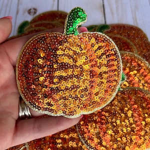 May include: Close-up of several pumpkin-shaped sequin patches. The patches are primarily orange and gold, with a green sequined stem. Each patch has a gold border. The patches are likely for crafting or embellishment.