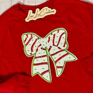 Pu&ograve; includere: Una felpa rossa con un grande fiocco decorativo. Il fiocco &egrave; bianco con strisce rosse e bordi verdi. L'etichetta in alto a destra recita "Lexi Ju Creations".
