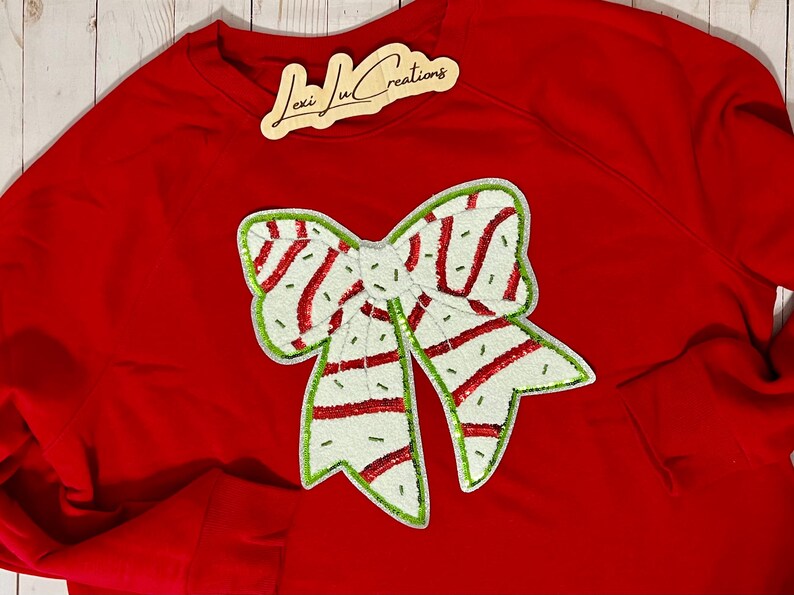 Pu&ograve; includere: Una felpa rossa con un grande fiocco decorativo. Il fiocco &egrave; bianco con strisce rosse e bordi verdi. L'etichetta in alto a destra recita "Lexi Ju Creations".