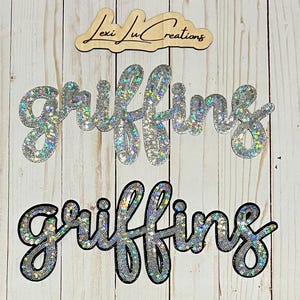 May include: Wooden sign with the text "Lexi Ju Creations" in a cursive font. Below are two silver glitter "Griffins" name cutouts, one with a black outline. The background is a light wood grain.