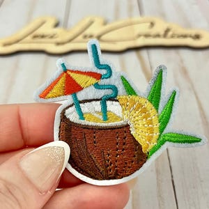 May include: Embroidered patch featuring a tropical cocktail in a coconut shell. The design includes a yellow and orange umbrella, a blue straw, and a pineapple slice. The coconut is brown with a white rim and a light blue border.