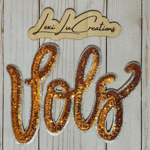 May include: A wooden sign with the text "Lexi Ju Creations" in a cursive font. Below, the word "fofo" is spelled out in large, orange sequins with a silver border, set against a light wood background.