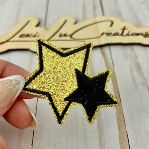 May include: A gold glitter star patch with a black outline and a smaller black star patch. The patches are held in a hand, with a wooden sign in the background that reads "Lexi Lu Creations".