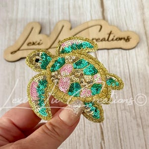 May include: A sea turtle patch featuring intricate embroidery and shimmering sequins in shades of emerald green, soft pink, and metallic gold. The turtle design has a detailed shell pattern and defined flippers, crafted with a gold-toned border.