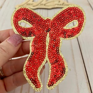 Könnte beinhalten: Eine rote Pailletten-Schleife mit einem goldglitzernden Rand. Die Schleife wird von einer Hand gehalten. Der Artikel ist ein dekorativer Gegenstand.