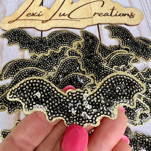 May include: A close-up of a pile of black and gold sequined bat patches. The bats are about 3 inches wide and have a gold glitter outline.