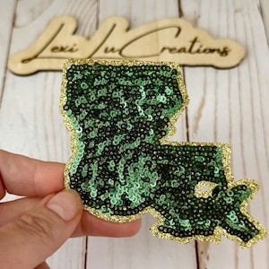 May include: A green sequined patch in the shape of the state of Louisiana, with a gold glitter border. The patch is held up in front of a wooden sign that says "Lexi Ju Creations".