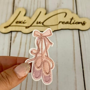 May include: Embroidered patch of a pair of pink ballet pointe shoes with a bow. The patch is on a white background with a light pink border. The wooden sign in the background reads "Lexi Lu Creations".