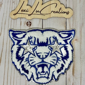 May include: A white tiger face with blue stripes and silver glitter stars for eyes. The tiger's mouth is open and its teeth are visible. The background is a light wood grain.