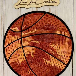 Può includere: Un design rotondo a forma di pallone da basket con paillettes arancioni e linee nere, che ricorda un vero pallone da basket. Il design è ambientato su uno sfondo di legno chiaro. Il testo "Lexi Lu Creations" è sopra il pallone.