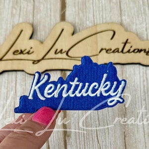 May include: A blue embroidered patch in the shape of the state of Kentucky with the word "Kentucky" in white script. The patch is held by a hand with pink nail polish. A wooden sign with the text "Lexi Lu Creations" is in the background.