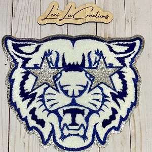 May include: A white glitter tiger head with blue details and silver glitter stars for eyes. The tiger has a large, open mouth with a blue "T" in the center.